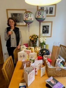 Joanna in the background raising a glass with two birthday balloons and a table full of birthday cards and flowers.