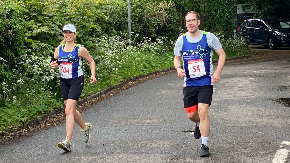 A female and male runner running alongside each other in a road race. Both are wearing Chilterns MS Centre running vests.