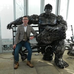 Man sitting next to a large robotic model of the Hulk.