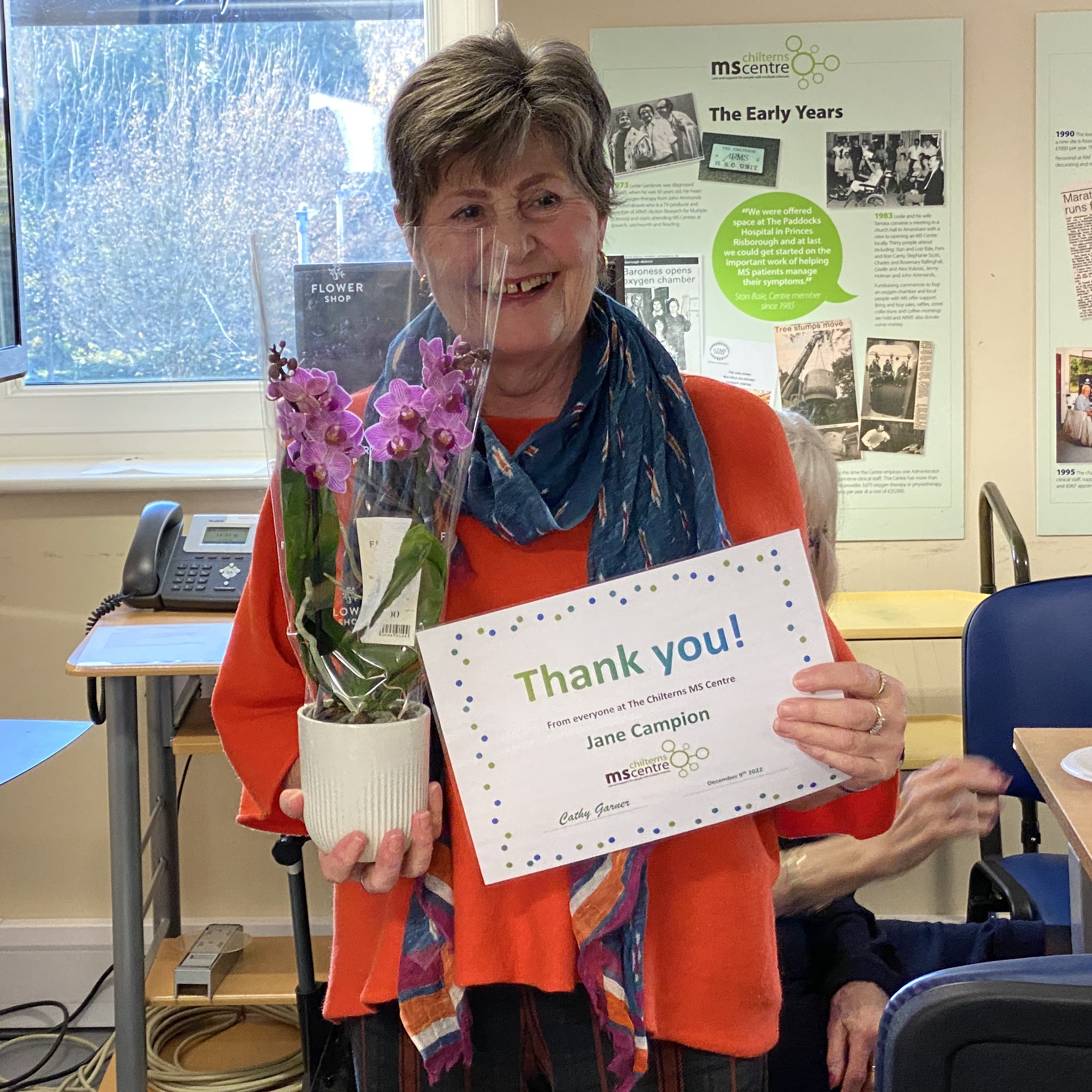 Smiling lady holding up a certificate saying thank you in one hand and a flower in the other.