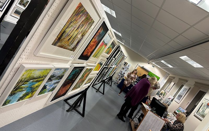 View of a busy art exhibition with paintings on the wall and on central frames.