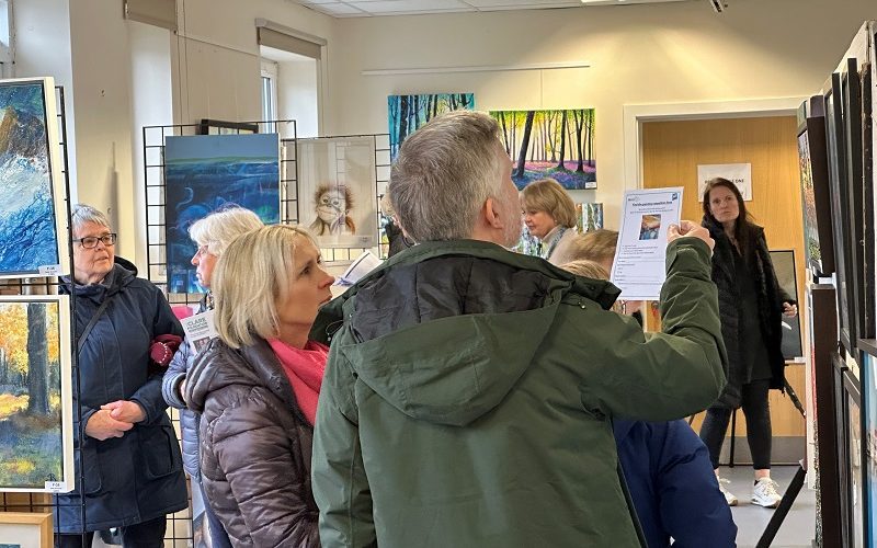 View of a busy art exhibition with paintings on the wall and on central frames.