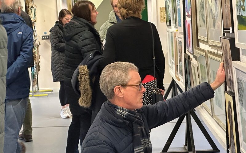 View of an art exhibition with a man crouching down looking intently at a painting.