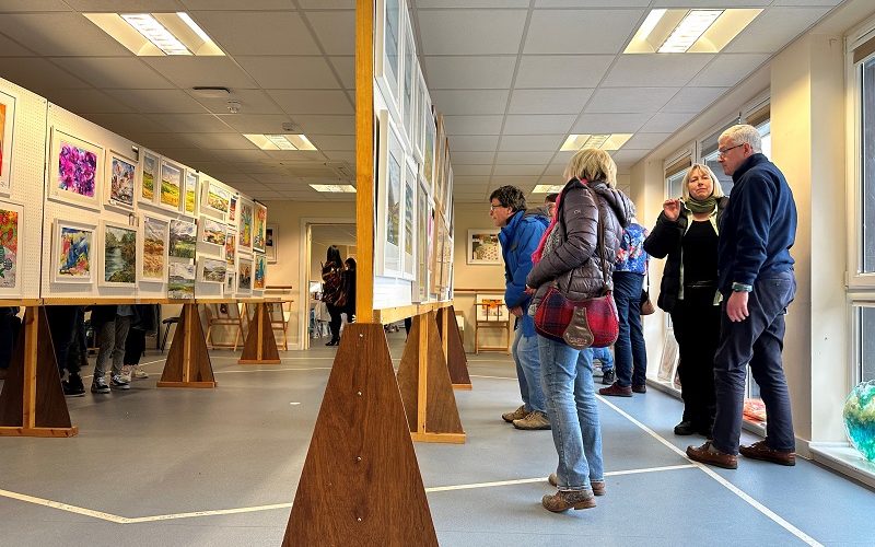 View of a busy art exhibition with paintings on the wall and on central frames.