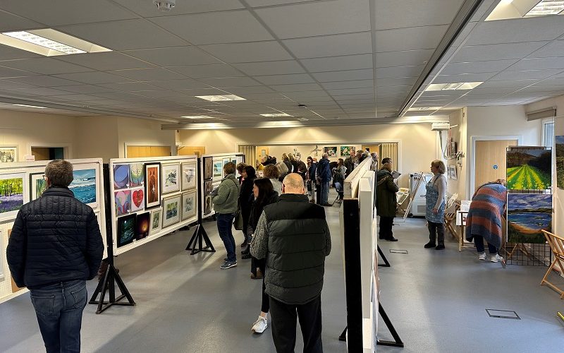 View of a busy art exhibition with paintings on the wall and on central frames.