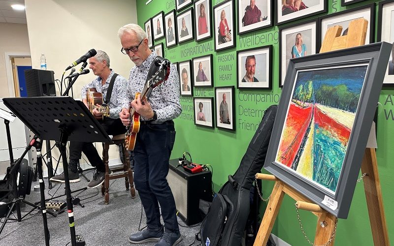 Two musicians playing guitars and singing into microphones. In the foreground is s painting of a countryside at sunset scene