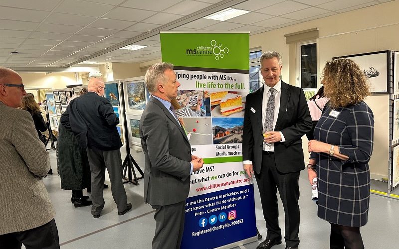 Two men and a woman standing in an art exhibition in front of banner promoting the Chilterns MS Centre