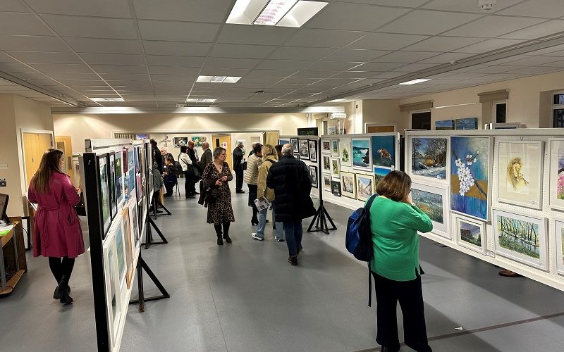 View of a busy art exhibition with paintings on the wall and on central frames.