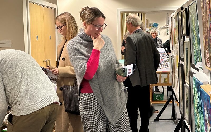 View of a busy art exhibition with paintings on the wall and on central frames.