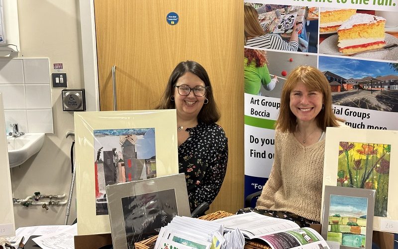 Two smiling women sitting behind a table with lots of greetings cards on.